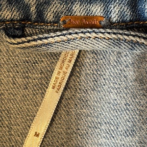 May include: Close-up of a denim jacket with a copper-coloured tag that reads "Free People". A white tag with the text "MADE IN MOROCCO FABRIQUE AU MAROC" is visible, along with the size "M".