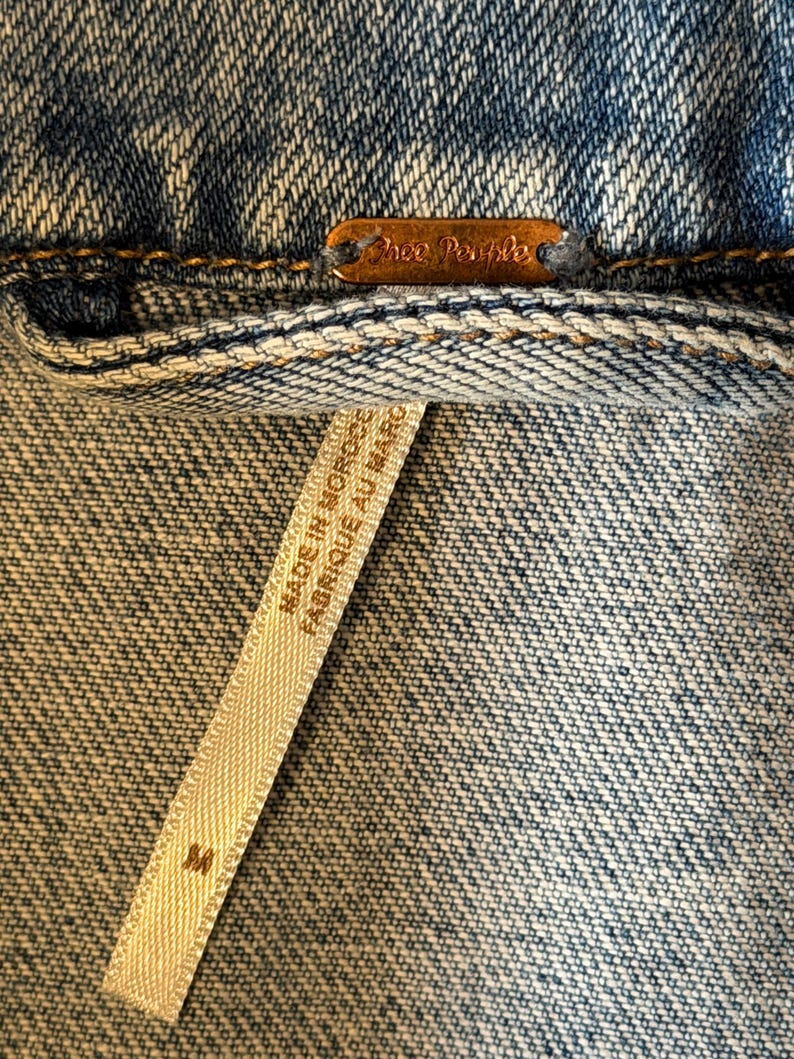 May include: Close-up of a denim jacket with a copper-coloured tag that reads "Free People". A white tag with the text "MADE IN MOROCCO FABRIQUE AU MAROC" is visible, along with the size "M".