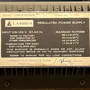 May include: A grey Lambda regulated power supply with black text. The label includes model, serial number, input voltage, and maximum ratings. The text also warns about surface temperature and ventilation.
