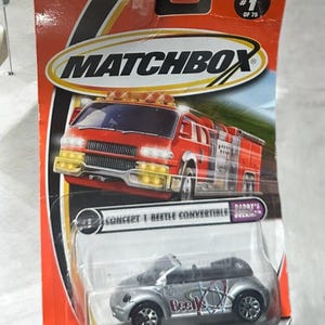 Puede incluir: Un coche de juguete Volkswagen Beetle descapotable plateado Matchbox con un logotipo "Beetle" en el lateral. El coche está en un blíster de plástico con fondo rojo y el logotipo de Matchbox. El embalaje también incluye un camión de bomberos rojo.