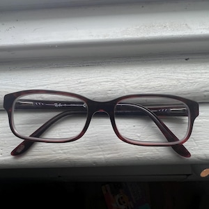 May include: A pair of rectangular eyeglasses with a dark burgundy frame and temples. The lenses are clear, and the glasses are resting on a white surface. The Ray-Ban logo is visible on the temple.