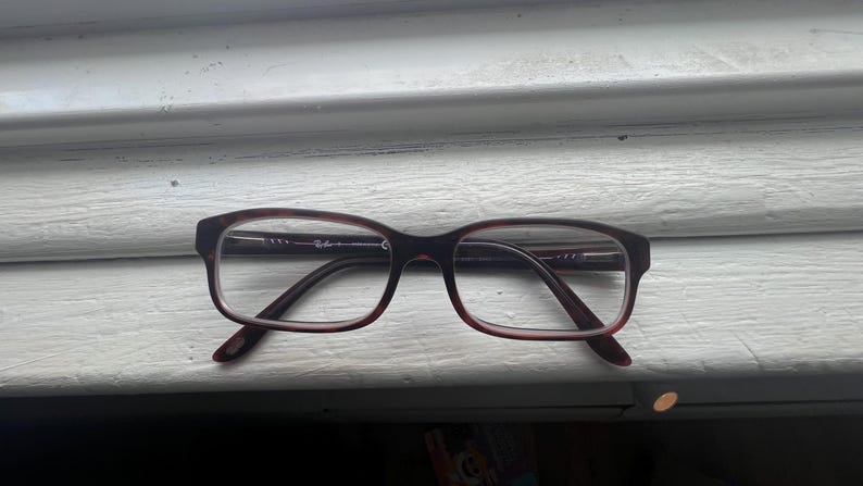 May include: A pair of rectangular eyeglasses with a dark burgundy frame and temples. The lenses are clear, and the glasses are resting on a white surface. The Ray-Ban logo is visible on the temple.
