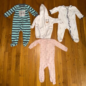 May include: Four baby sleepwear items are displayed on a wooden surface. There is a blue and gray striped footed sleeper with a bear face, a white hooded robe with a zebra, a pink footed sleeper, and a white dotted sleeper with navy trim.