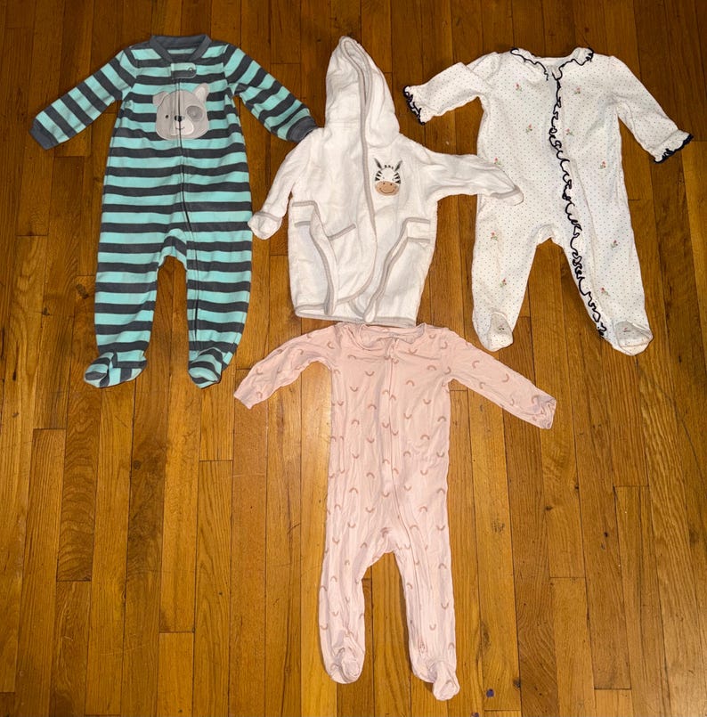 May include: Four baby sleepwear items are displayed on a wooden surface. There is a blue and gray striped footed sleeper with a bear face, a white hooded robe with a zebra, a pink footed sleeper, and a white dotted sleeper with navy trim.