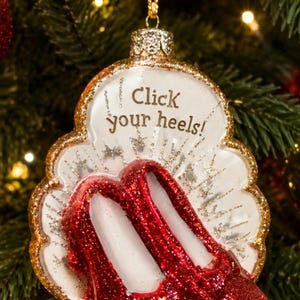 May include: A white, scalloped-edge Christmas ornament featuring a pair of glittery red shoes. The ornament has gold glitter trim and the text "Click your heels!" printed in gold. It hangs from a gold string and includes a gold metal cap.