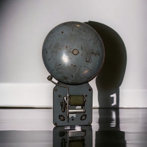 May include: A vintage gray metal bell with a rounded top and a rectangular base. The bell has a small lever and a coil visible on the front. The bell is likely used for signaling or alarms.