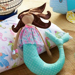 May include: A handmade mermaid doll with brown hair and a floral top. The doll has a turquoise tail with white polka dots and is resting on a pillow with seahorse illustrations. A pink bow accents the doll's wrist.