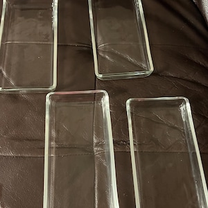 May include: Five clear, rectangular glass trays. Each tray has a slightly raised edge and a simple, minimalist design. The trays are arranged on a brown surface.