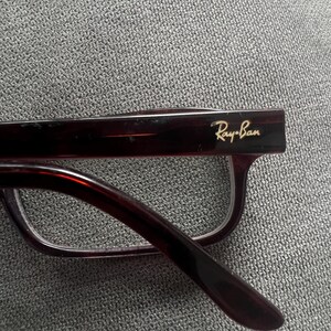 May include: Close-up of Ray-Ban eyeglasses with a dark burgundy frame. The Ray-Ban logo is visible in gold lettering on the temple. The glasses have a classic rectangular shape and are set against a gray textured background.