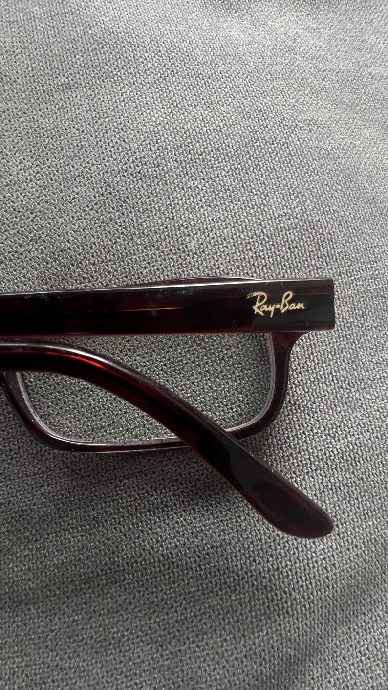 May include: Close-up of Ray-Ban eyeglasses with a dark burgundy frame. The Ray-Ban logo is visible in gold lettering on the temple. The glasses have a classic rectangular shape and are set against a gray textured background.