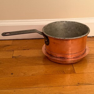 May include: A copper saucepan with a long, dark metal handle. The pot has a rounded base and a slightly flared rim. It sits on a copper base. The interior of the pot shows signs of use. The pan is likely used for cooking.