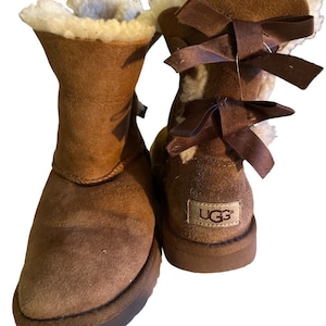 May include: A pair of brown suede boots with a light brown interior and a dark brown sole. The boots have a bow detail on the back and the brand name "UGG" is visible on the heel.