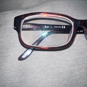 May include: Brown and black Ray-Ban eyeglasses with a rectangular frame. The glasses have clear lenses and the Ray-Ban logo is visible on the frame. The temples are dark brown and the glasses are resting on a gray surface.