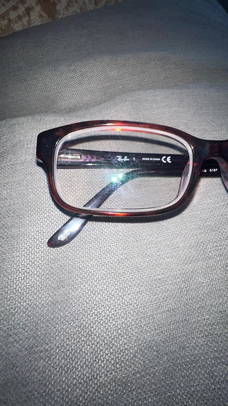 May include: Brown and black Ray-Ban eyeglasses with a rectangular frame. The glasses have clear lenses and the Ray-Ban logo is visible on the frame. The temples are dark brown and the glasses are resting on a gray surface.