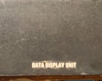Puede incluir: Una unidad de visualización de datos de color gris oscuro con dos asas metálicas y una etiqueta blanca con texto y un interruptor. El texto "DATA DISPLAY UNIT" está impreso en la parte delantera.