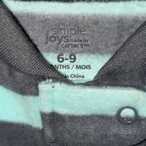 May include: Close-up of a baby's garment with horizontal stripes in shades of gray and teal. The label reads "simple joys made by carter's" and indicates the size as 6-9 months. The garment has a snap closure.