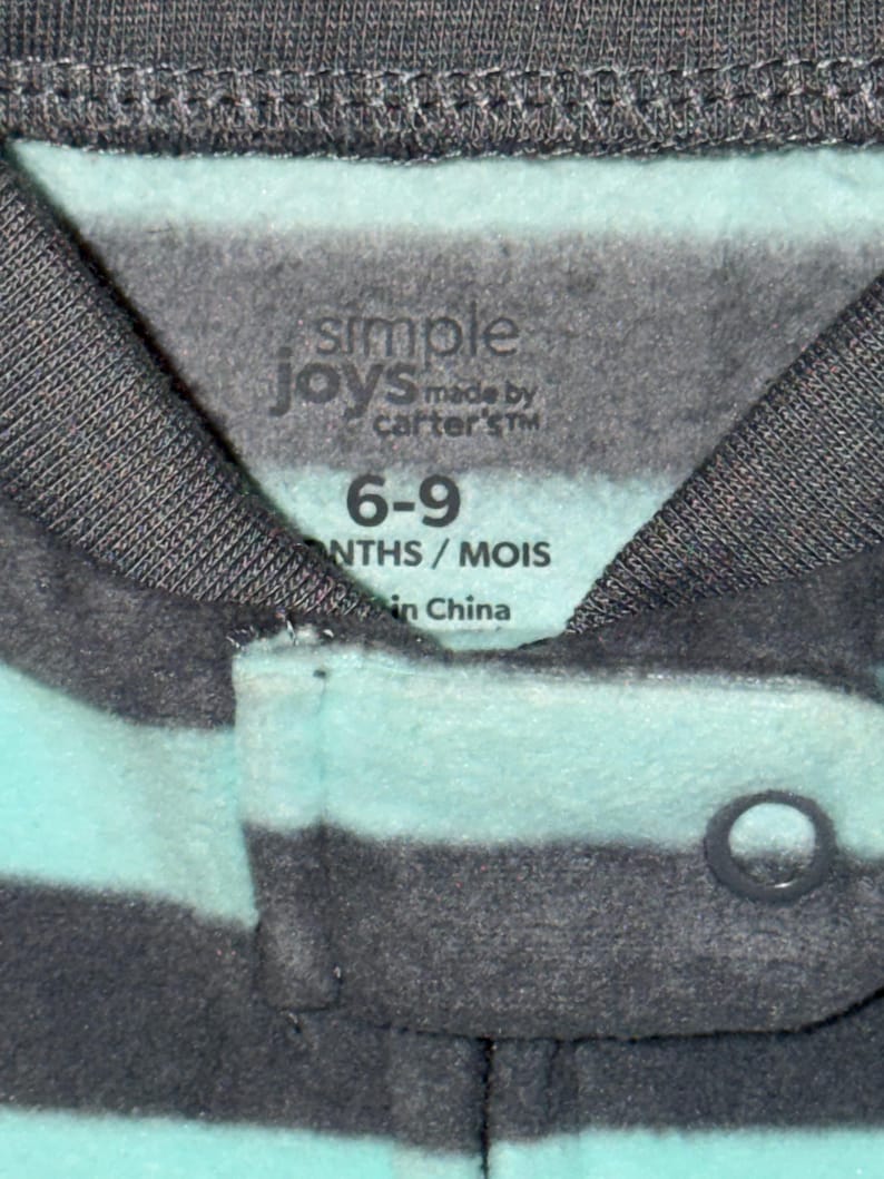 May include: Close-up of a baby's garment with horizontal stripes in shades of gray and teal. The label reads "simple joys made by carter's" and indicates the size as 6-9 months. The garment has a snap closure.