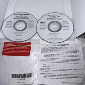Może przedstawiać: Dwa dyski CD-ROM i papierowa instrukcja obsługi systemu odzyskiwania danych komputera. Dyski są oznaczone jako "Product Recovery CD-ROM for IBM ThinkPad A30m" i "Product Recovery CD-ROM for IBM ThinkPad A30m". Instrukcja obsługi ma tytuł "Instructions for using the Recovery CD".