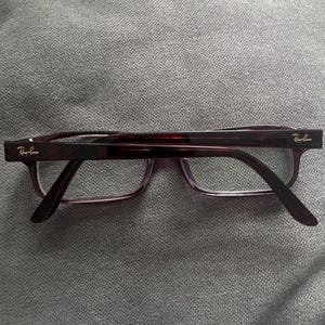 May include: Ray-Ban eyeglasses with a dark burgundy frame and clear lenses. The temples have the Ray-Ban logo in gold. The glasses are rectangular and sit on a gray surface.