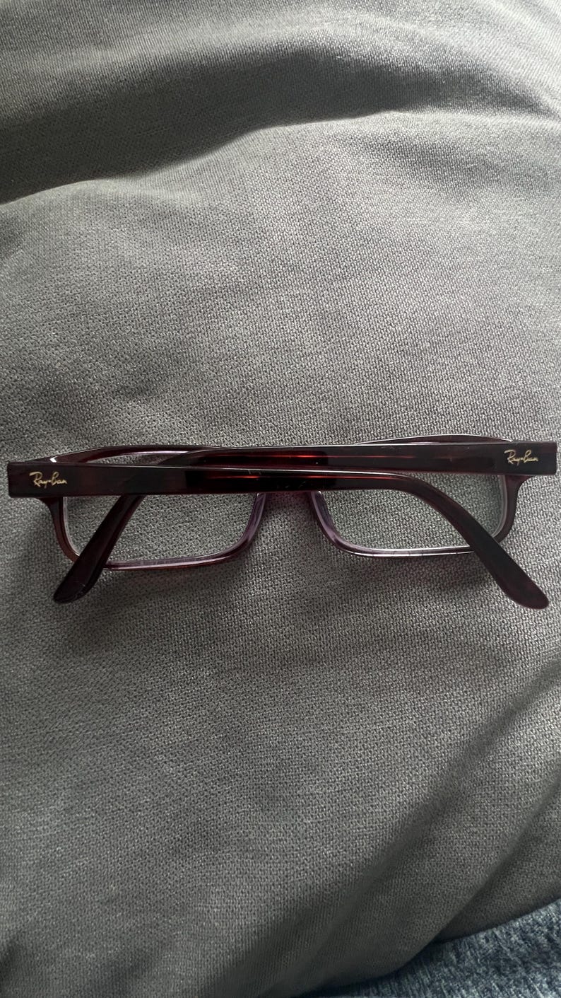 May include: Ray-Ban eyeglasses with a dark burgundy frame and clear lenses. The temples have the Ray-Ban logo in gold. The glasses are rectangular and sit on a gray surface.