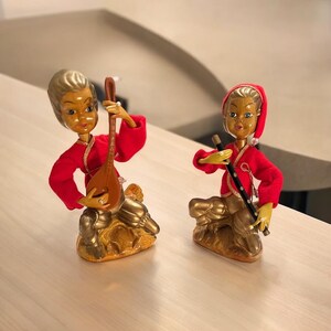 May include: Two small figurines of people playing musical instruments. The figurine on the left is playing a lute and is wearing a red and gold outfit. The figurine on the right is playing a flute and is wearing a red and gold outfit with a red hat.