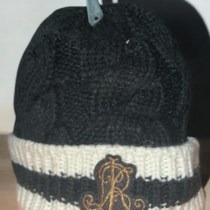 May include: A black knit beanie with a white and black striped brim. The beanie features a gold embroidered emblem with the letter "R". The hat is displayed with a clear plastic hanger attached.