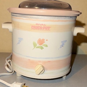 Puede incluir: Una olla de cocción lenta Rival Crock-Pot blanca con tapa transparente y detalles en rosa. La olla de cocción lenta tiene un diseño floral y el texto "RIVAL CROCK-POT STONEWARE SLOW COOKER". Tiene un dial de tres posiciones y un cable de alimentación.