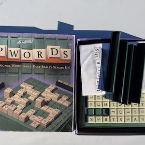 May include: The Upwords board game box is open, revealing the game board, letter tiles, and game pieces. The box lid displays the game's name and tagline. The game board is green and the letter tiles are beige.