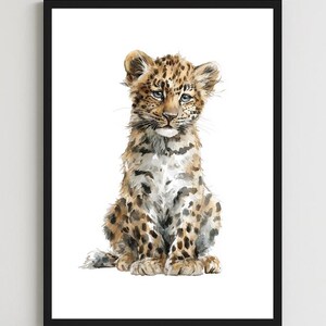 May include: A framed watercolor print of a sitting leopard cub. The artwork features the cub with a spotted coat in shades of brown and tan, with blue eyes. The print is in a black frame and is displayed on a white wall.