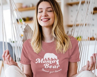 May include: A woman wearing a pink t-shirt with a white graphic of a mama bear and a baby bear. The text on the shirt reads "Mama Bear Sweet yet fierce".