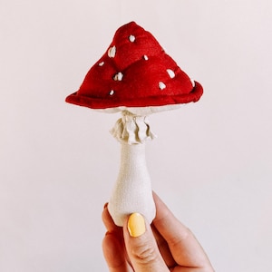 May include: A red and white fabric mushroom toy with white polka dots. The mushroom is held in a hand with yellow nail polish.