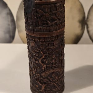 Könnte beinhalten: Ein zylindrischer, dunkelbrauner, geschnitzter Holzbehälter. Der Behälter weist detaillierte Reliefschnitzereien einer Landschaft mit Figuren, Gebäuden und Brücken auf. Ober- und Unterseite sind mit dekorativen Schnitzereien versehen.