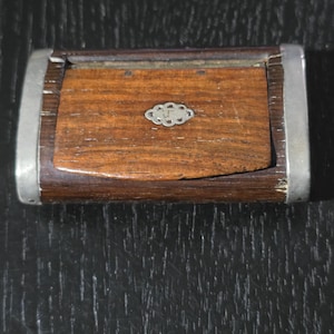 May include: A rectangular wooden box with a hinged lid and metal trim. The box has a dark brown finish with a small, decorative silver-colored emblem on the lid. The edges are reinforced with silver metal.