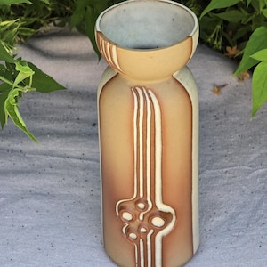 May include: A cylindrical ceramic vase featuring a flared rim and a smooth, matte finish in shades of tan and brown. The exterior is decorated with a vertical, raised white relief pattern consisting of parallel lines and circular shapes.