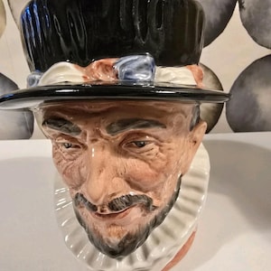 Puede incluir: Una taza de cerámica con forma de cabeza de hombre, con un sombrero de copa negro y un cuello con volantes blanco. La cara está detallada con bigote y barba, pintados en tonos marrones y rosas.