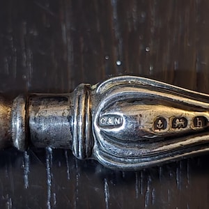 May include: Close-up of a silver utensil handle with ornate detailing. The handle features the letters "C&N" within a rectangular cartouche, along with several hallmarks. The handle is likely part of a knife or fork.