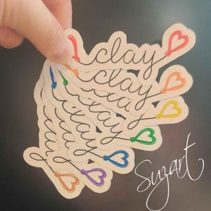 May include: A stack of white stickers with the word "clay" in black cursive, each with a colorful heart design. The hearts are in various colors, including red, orange, yellow, green, blue, and purple. The artist's signature "Suzart" is in white cursive.