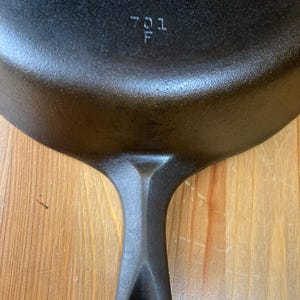 May include: A black cast iron skillet featuring a textured surface and a handle. The number 701 and the letter F are clearly embossed on the top of the pan near the handle connection point, showing a dark metallic finish and sturdy construction.