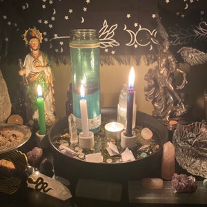 Puede incluir: Una bandeja negra con velas, cristales y otros artículos espirituales. Hay una estatua de un santo, un frasco de vidrio con líquido azul y un tazón pequeño con incienso. La bandeja está sobre una mesa con un fondo oscuro.