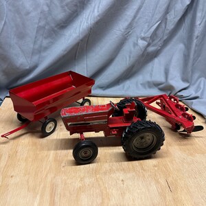May include: Red toy tractor with a wagon and a plow. The tractor has the word "INTERNATIONAL" printed on the side.