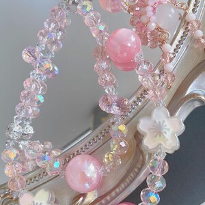 May include: A selection of pink beaded accessories, including a keychain and bracelets. The items feature iridescent crystals, large pink spheres, and white flower-shaped charms. The accessories are displayed against a reflective surface.