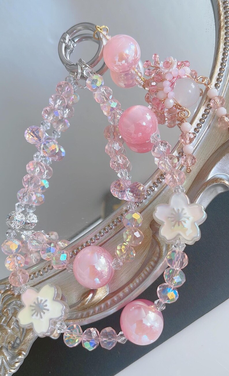May include: A selection of pink beaded accessories, including a keychain and bracelets. The items feature iridescent crystals, large pink spheres, and white flower-shaped charms. The accessories are displayed against a reflective surface.