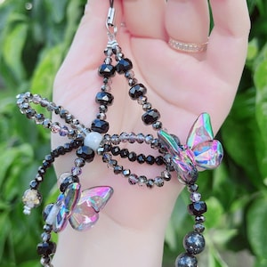 May include: A beaded phone charm featuring a butterfly design. The charm has black and iridescent beads, with two iridescent butterfly charms. The charm is held by a silver-colored clasp.