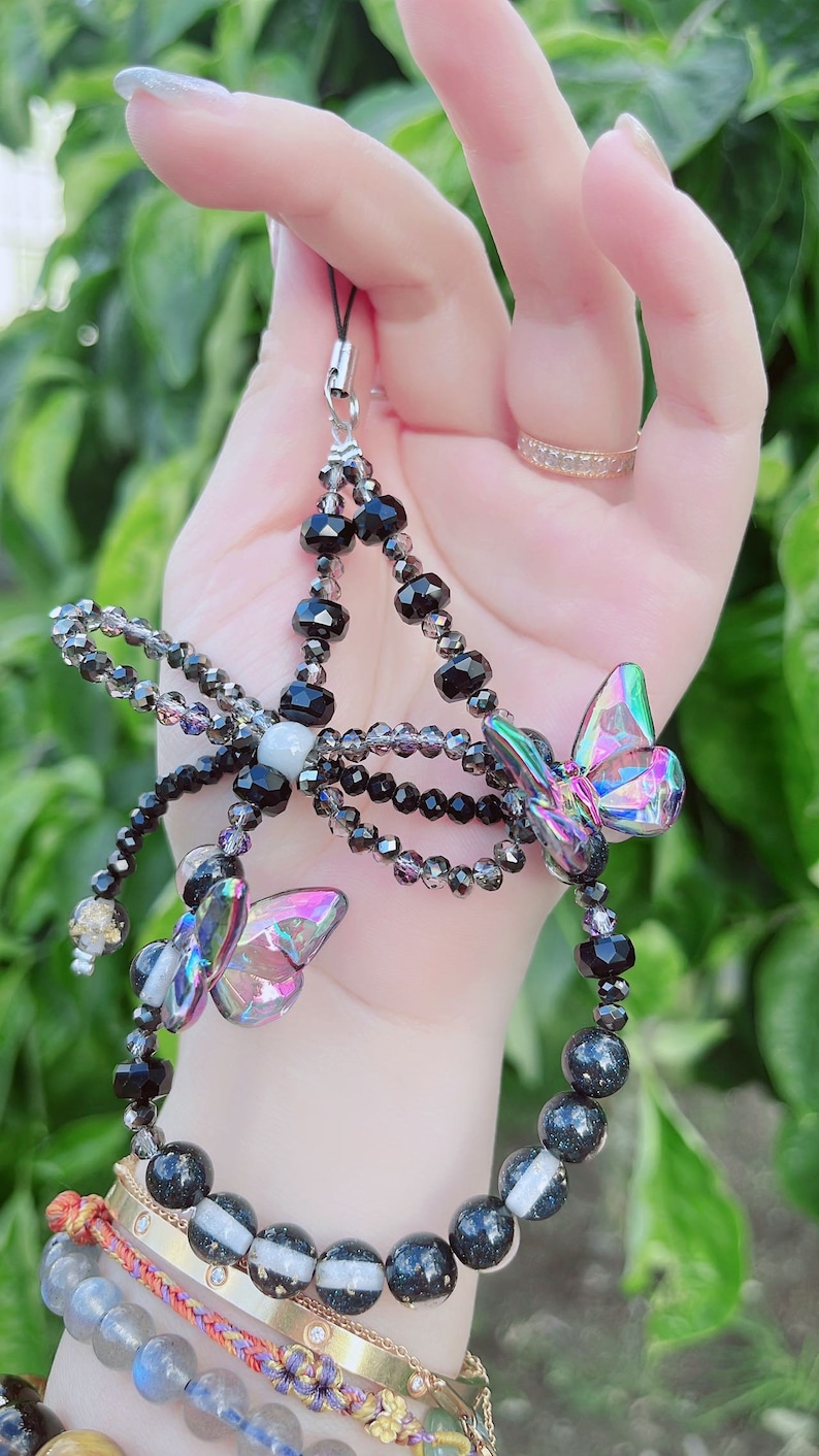 May include: A beaded phone charm featuring a butterfly design. The charm has black and iridescent beads, with two iridescent butterfly charms. The charm is held by a silver-colored clasp.