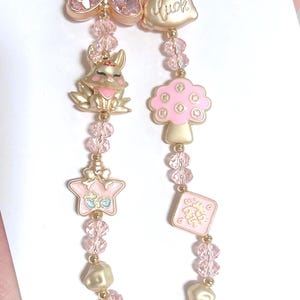 May include: A gold-coloured keychain with pink and gold charms. Charms include a bow, a mushroom, a star, a rabbit, and a small gold bar with the text "FINE GOLD 9999 1000."