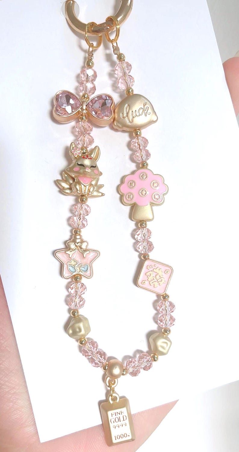 May include: A gold-coloured keychain with pink and gold charms. Charms include a bow, a mushroom, a star, a rabbit, and a small gold bar with the text "FINE GOLD 9999 1000."