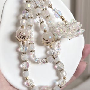 May include: A beaded keychain with a gold-tone clasp. The keychain features strands of white, iridescent, and clear beads, with gold accents and a decorative flower charm. The beads are various shapes and sizes, creating a layered effect.