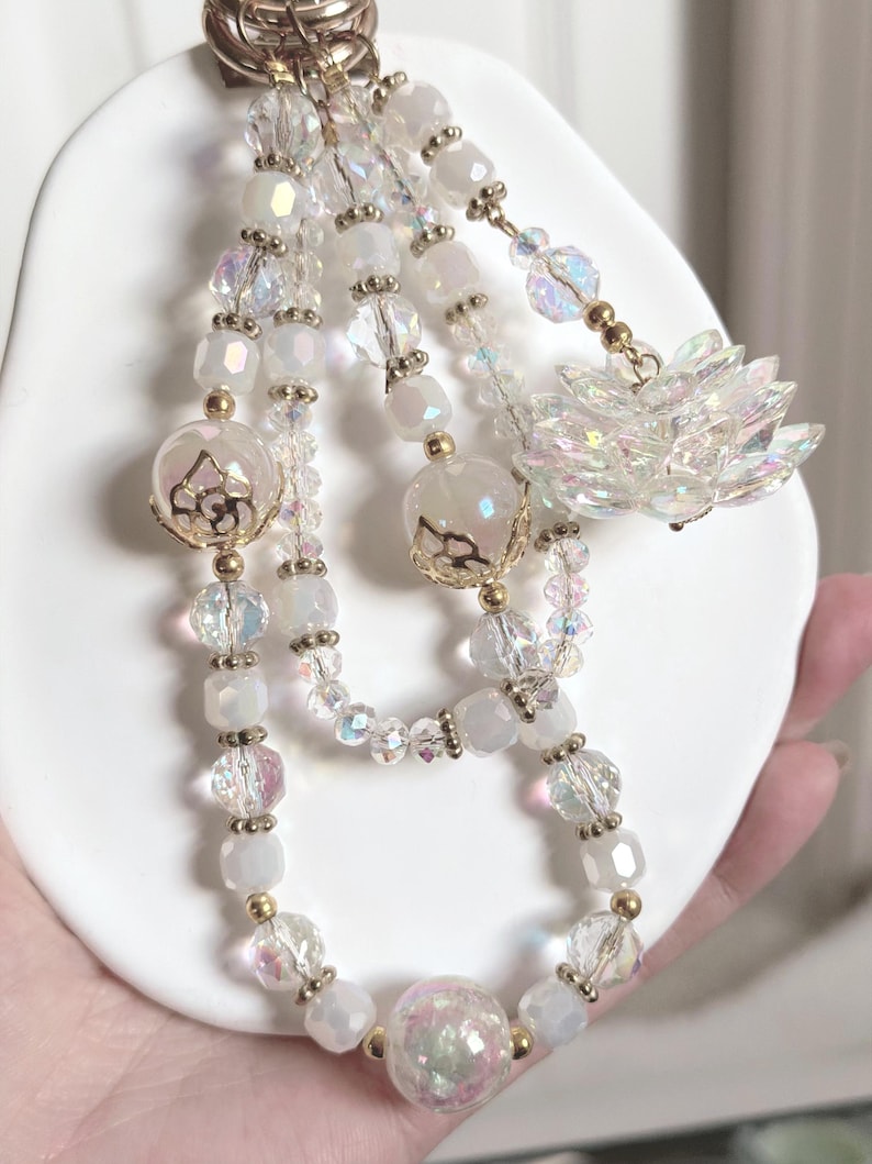 May include: A beaded keychain with a gold-tone clasp. The keychain features strands of white, iridescent, and clear beads, with gold accents and a decorative flower charm. The beads are various shapes and sizes, creating a layered effect.