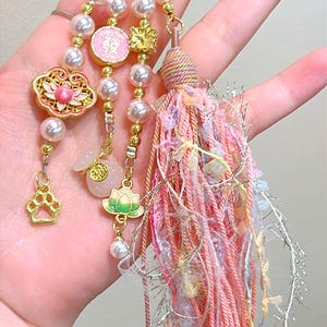 May include: A decorative keychain with a gold ring, featuring pearl beads, pink and gold charms, and a tassel. Charms include a lotus flower, paw print, and a pink circular charm with Chinese characters.