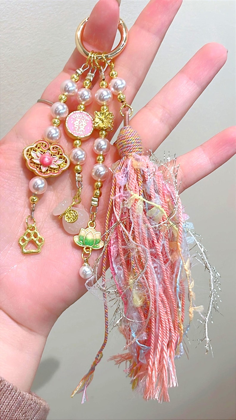 May include: A decorative keychain with a gold ring, featuring pearl beads, pink and gold charms, and a tassel. Charms include a lotus flower, paw print, and a pink circular charm with Chinese characters.